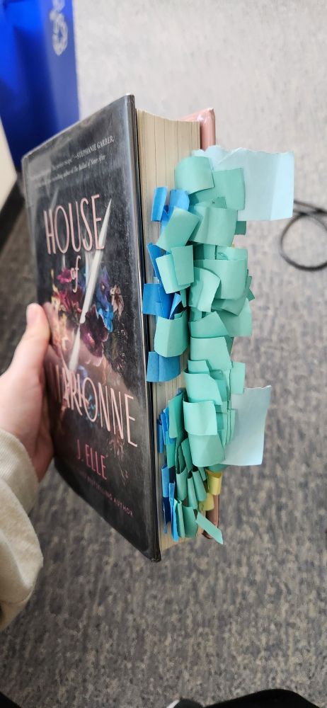 A copy of HOUSE OF MARIONNE by J. Elle is being held angled so you can see the cover and the pages of the book. The pages are overflowing with blue and teal and a few yellow small sticky notes. 