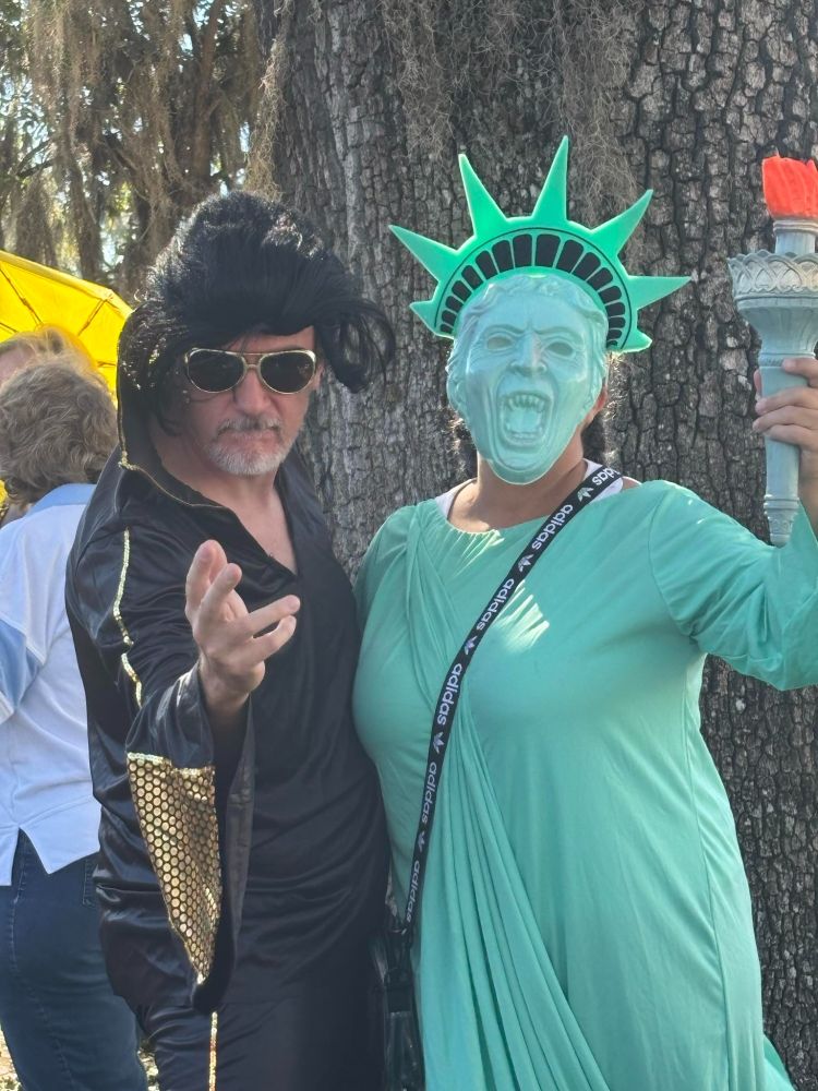 Two protesters: man in black Elvis-style costume, person in Statue of Liberty mask with screaming expression and green dress.