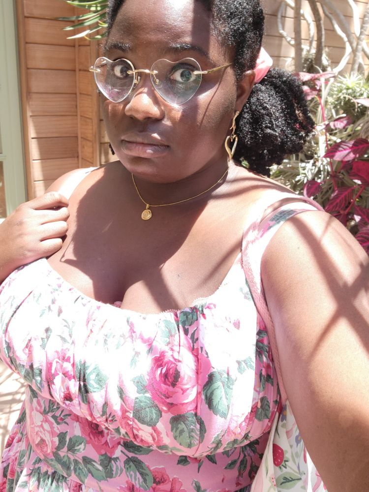 A black woman in a pink floral dress wearing heart shaped glasses, gazing pensively at the camera.