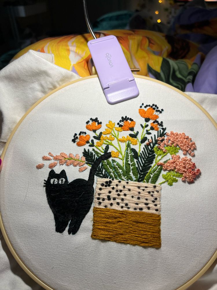 A black cat is next to a roped planter that has green plants and flowers that are coral, yellow, and orange