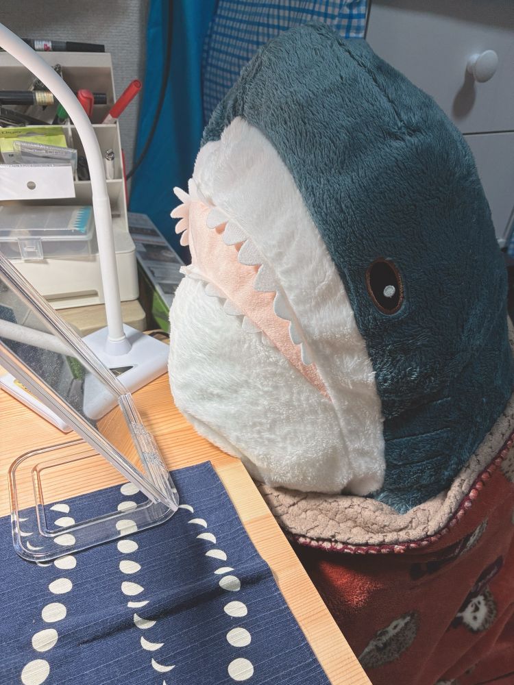 テーブルにもたれながら卓上の鏡の方を見ている大きなサメのぬいぐるみ
