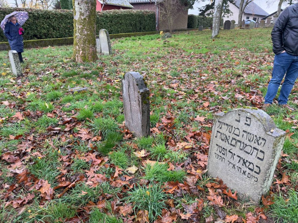 A Jewish cemetery on a rainy day showing old gravestones with Hebrew inscriptions amid fallen autumn leaves and grass. Two people can be partially seen in the background, one holding an umbrella.