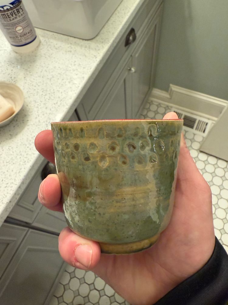 A hand is holding a small ceramic cup with a greenish glaze and a textured pattern around the upper half. The cup features a shiny finish with subtle variations in color, ranging from olive to mint green. The hand holding the cup is positioned over a white and gray tile floor with a hexagonal pattern and a light-colored cabinet in the background. The cup's organic shape and handmade appearance add to its rustic charm.￼