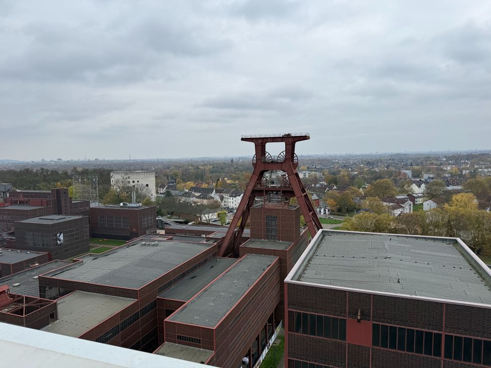An elevated view of the Zollverein Coal Mine complex in Essen, showing the iconic red-brown mining headframe tower and industrial buildings against an overcast sky and urban landscape.