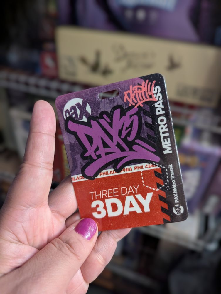 I'm holding in my left hand my 3-day Pax Unplugged 2025 badge.

My manicure is still holding up; I got it last weekend at Beauty Bar in the East Village, where they have a late-night manicurist on site at their bar where you can buy a drink + manicure special.

Blurred background is one of my board game shelves, full-to-bursting. The most prominent blurred title is the beige fat box that is Darwin's Journey.