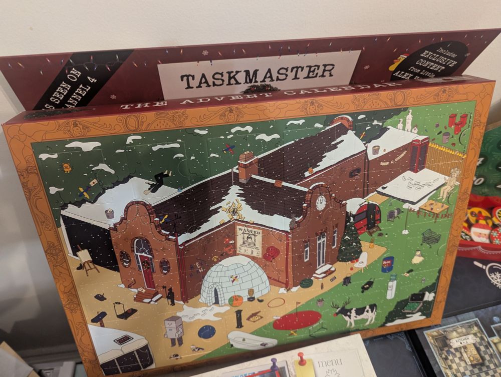 Taskmaster Advent Calendar. The box itself is the calendar. It's a 3/4 illustration of the Taskmaster house and grounds, populated with various references to Taskmaster lore. The roof has snow on it. There are unopened doors all over the illustration and box, which are opened in a specific order that is only known if you complete the tasks and puzzles in the correct order. Each puzzle's answer reveals which door to open next. The object is to find Little Alex Horne over 24 "days". 