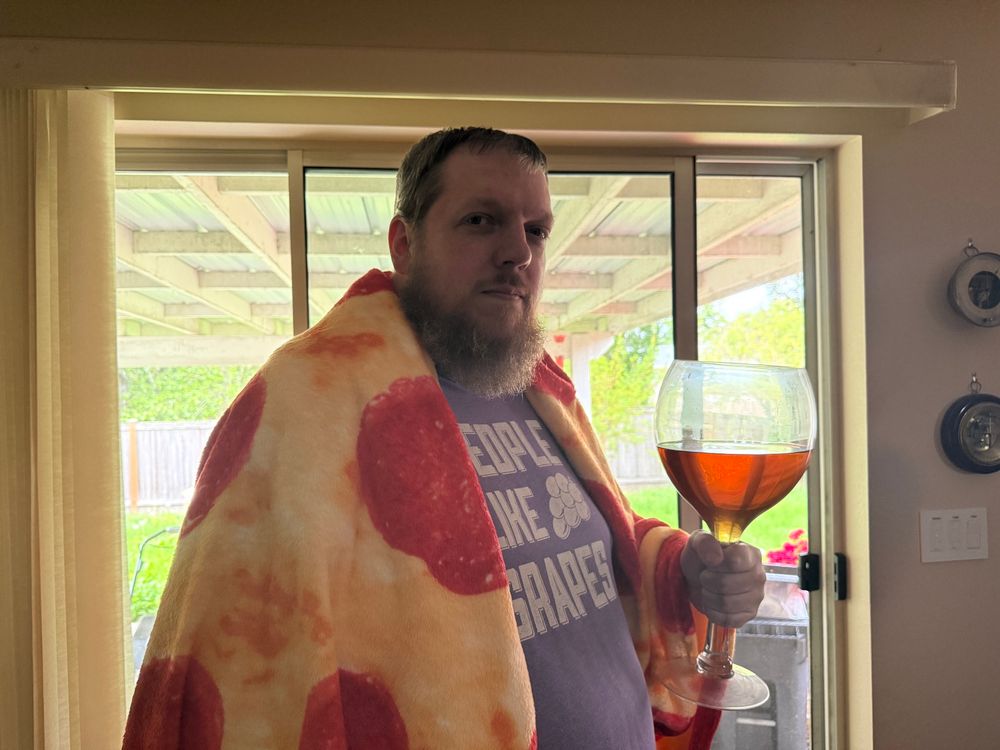 A man with a pizza blanket draped over his shoulders holding an oversized wine glass with an amber liquid in it