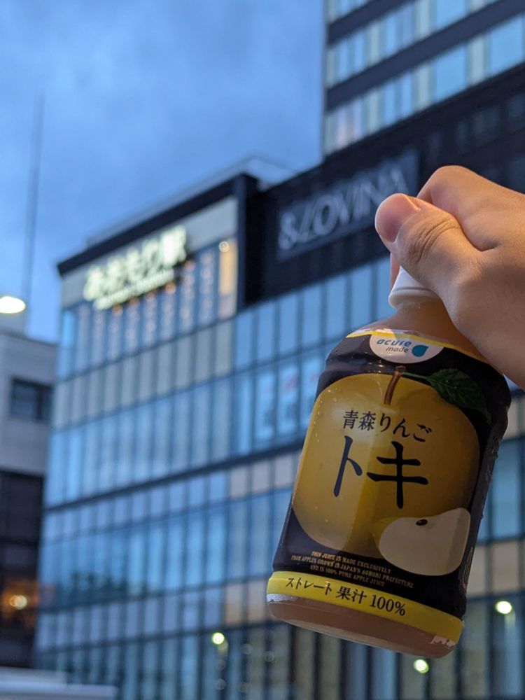 青森駅を背景に「青森りんご トキ」のラベルが巻かれたペットボトルが掲げられている写真。