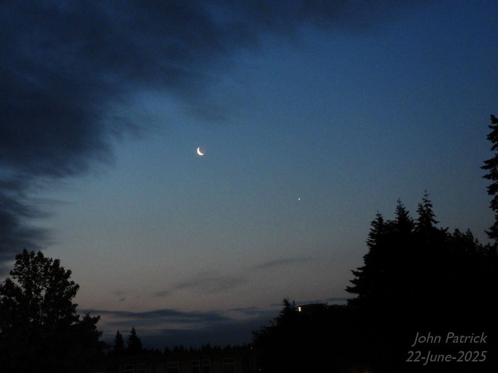 Photo: The moon and the planet Venus close together in the pre-dawn sky.  In the foreground are the silhouettes of trees that mirror the clouds rising from the lower left to the upper right.  Also in silhouette is a low apartment building with a single light on directly below Venus. Covering the sky from the lower left in a curving angle to the upper right are tendrils of clouds.  