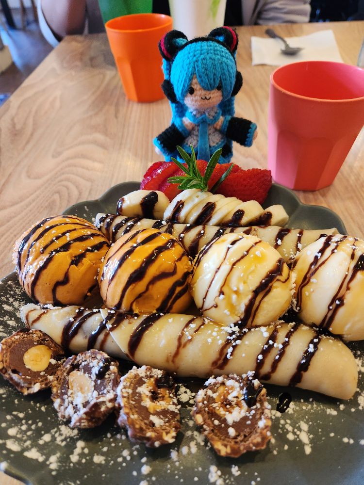 A plate of forrer roches cut in half and two rows of rolled up crepes, with four scoops of ice cream in between them. In the far back there are sliced bananas and strawberries; the entire plate has a drizzle of chocolate and powdered sugar. Amigurumi Hatsune Miku doll stands in the back by the plate.