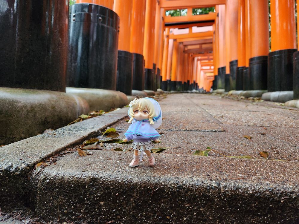 Nendoroid doll of Lumine with a little rain hoodie. She's walking through the thousand torii gates in Kyoto, looking up at them with a gentle smile