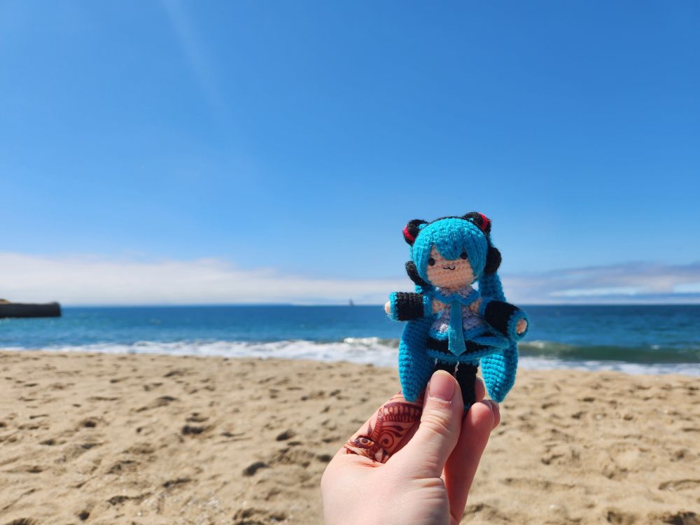 At the beach with Amigurumi Hatsune Miku doll held up in front of the shoreline. The blue of the ocean is similar to the blue of her hair.