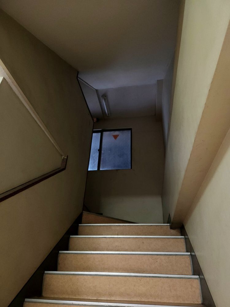 Steep stairs going up and around a corridor with very bland walls and no light at the top. At the top corner railing, a small fuwapuchi Airi plushie is seen poking out of the darkness. 
