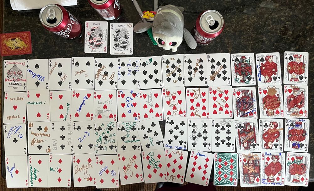 A deck of playing cards being watched over by a Duskull plushie. Each card is signed by a different SGDQ 2025 attendee. 