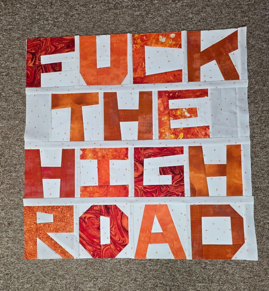 A quilt with orange letters on a white background spelling "fuck the high road".