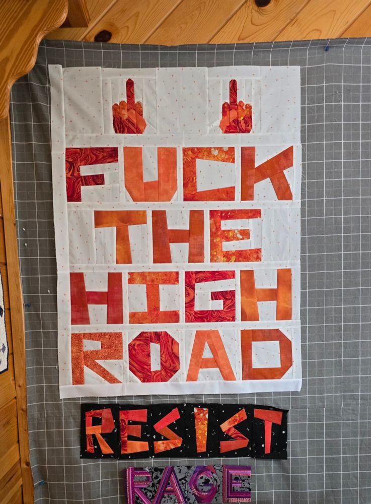 In fabric, two middle fingers and the words Fuck The High Road in orange ona white background.  Below, the words Resist and Rage also in fabric.