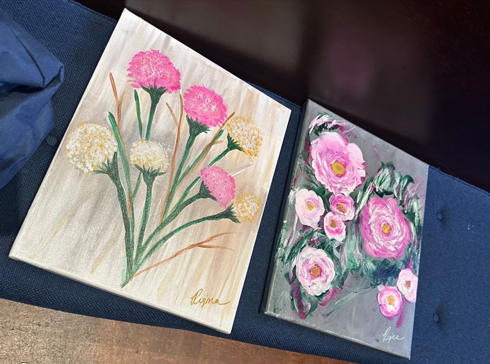 Two floral paintings side by side on a bench with blue cushions. The painting on the left has a tan abstract background with pink and gold carnations. The painting on the right has a gray abstract background with pink and white peonies. 