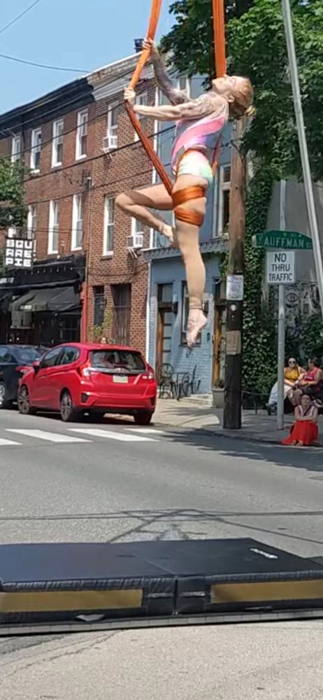 Woman in an orange sling, wrapped up vertically to a cupid position. Like all the other photos, there is a crash mat. 