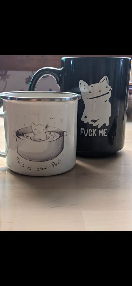On a blonde with table sits two mugs. In the foreground a small white enamel mug and in the background eats all black ceramic bug. On the small white mug is a picture of a soup pot with a little annoyed cat sitting in it with fluid and vegetables. It is of our cat named Puddle Cat so, it says "this is my Stu-pud mug" and the other is a very derpy, almost like ditto from pokémon, drawing of a white cat and under it says, "Fork me" but explicit. 