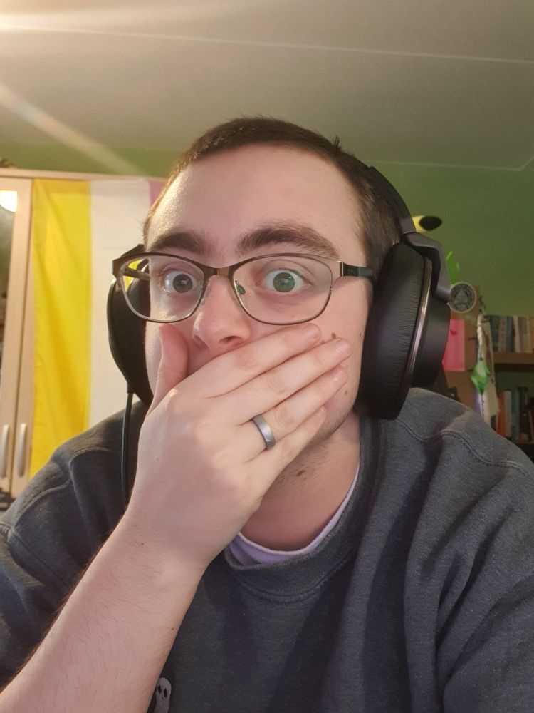 a selfie of Artie looking shocked and covering their mouth with their hand. They are wearing a grey jumper, large black headphones and glasses, with short buzzcut brown hair