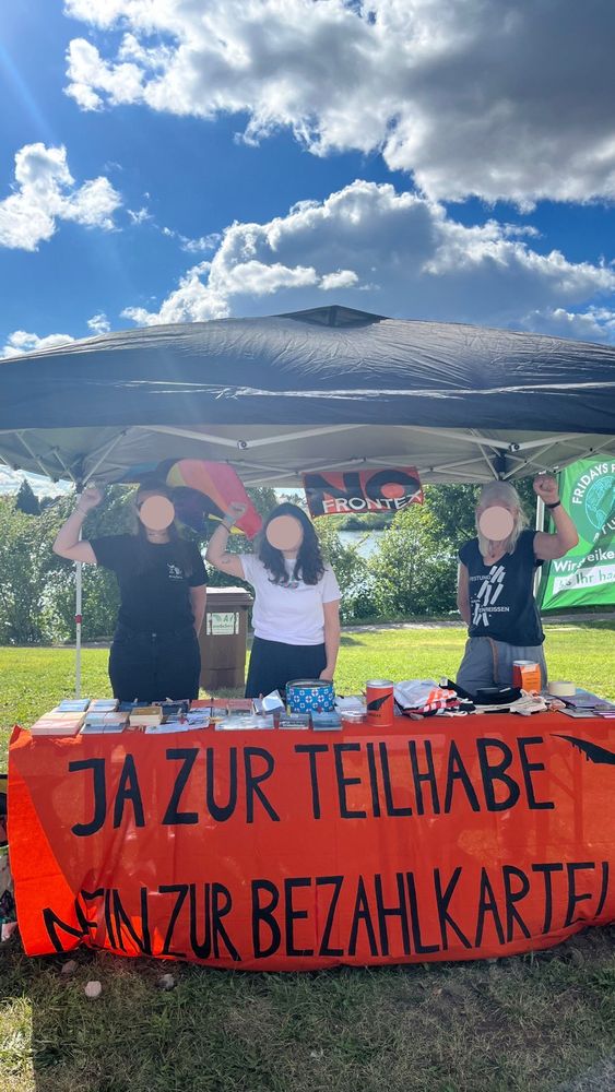 Unter einem Pavillon befindet sich ein Biertisch auf dem Infomaterial verschiedener NGOs wie z.B. Seebrücke, Flüchtlingsrat Baden - Württemberg und medico international ausgelegt ist. Dazu Buttons, T-Shirts, Beutel bedruckt mit z.B. „Festung EU einreißen“ oder „No Border“. Zusätzlich hängt an der Vorderseite des Tisches ein Transparent: auf orangem Stoff steht in schwarzen Blockbuchstaben: „Ja zur Teilhabe, nein zur Bezahlkarte!“ In der der unteren linken und der oberen rechten Ecke je ein kleine Schwarze Brücke. Rechts am Rand, unterhalb der Brücke, auch in schwarzen Blockbuchstaben „Seebrücke MA“ Hinter dem Tisch stehen 3 Menschen der Seebrücke MA und HD, die Gesichter verdeckt, die Fäuste oben. Hinten am Pavillon innen angemacht (also gut von vorne zu sehen) die Pride Flag mit der Ausschrift „Seebrücke Heidelberg“ und ein „No Frontex“ Transpi. Oranges Transpi auf dem im Hintergrund schwarz geschrieben, ganz groß (das ganze Transpi ausfüllend) „No“ steht und im Vordergrund, quer über das untere fünftel in weißer Schrift „Frontex“. Alles mit Blockbuchstaben geschrieben. Im Hintergrund Rasen,Bäume / Bäumchen, ein blauer Himmel mit einigen Wolken und rechts weht ein Teil der „Fridays for future“ Flagge ins Bild.