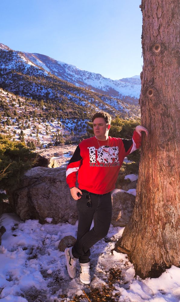 a picture of me leaning against a tree standing in snow with a mountain backdrop. I'm wearing a red 101 Dalmatians sweater, black jeans and white 101 Dalmatians high top Converse. the look is rounded out by a pair of sunglasses hanging from my waistband and a couple of silver necklaces