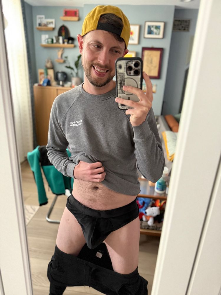 Mirror selfie of me in a yellow corduroy cap on backwards, grey sweatshirt, velvety black briefs, and black sweatpants pulled down a bit 