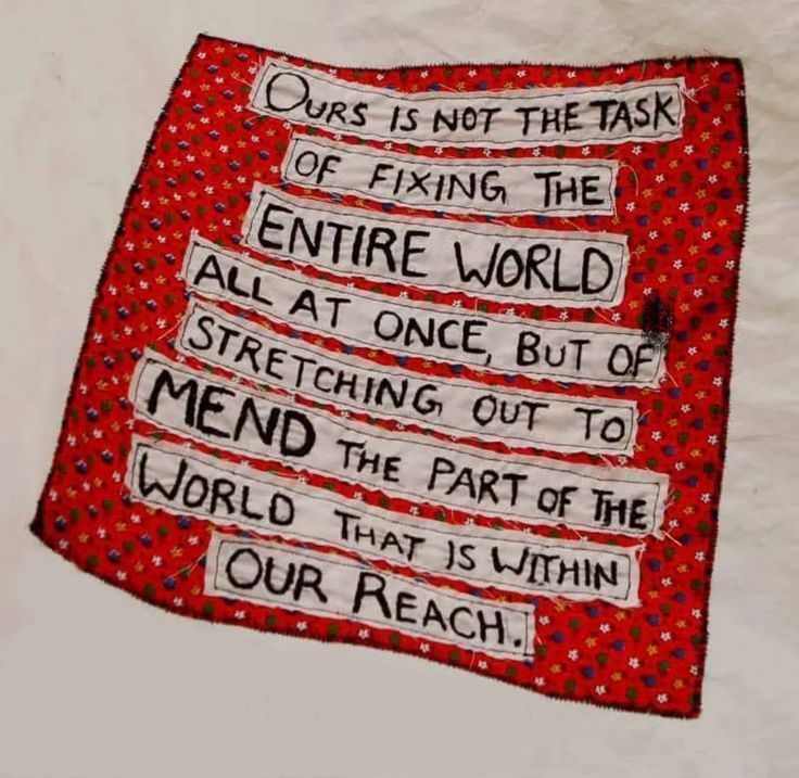 A hand-stitched fabric square with a red floral background featuring an embroidered quote in black and white text. The quote reads: “Ours is not the task of fixing the entire world all at once, but of stretching out to mend the part of the world that is within our reach!”
