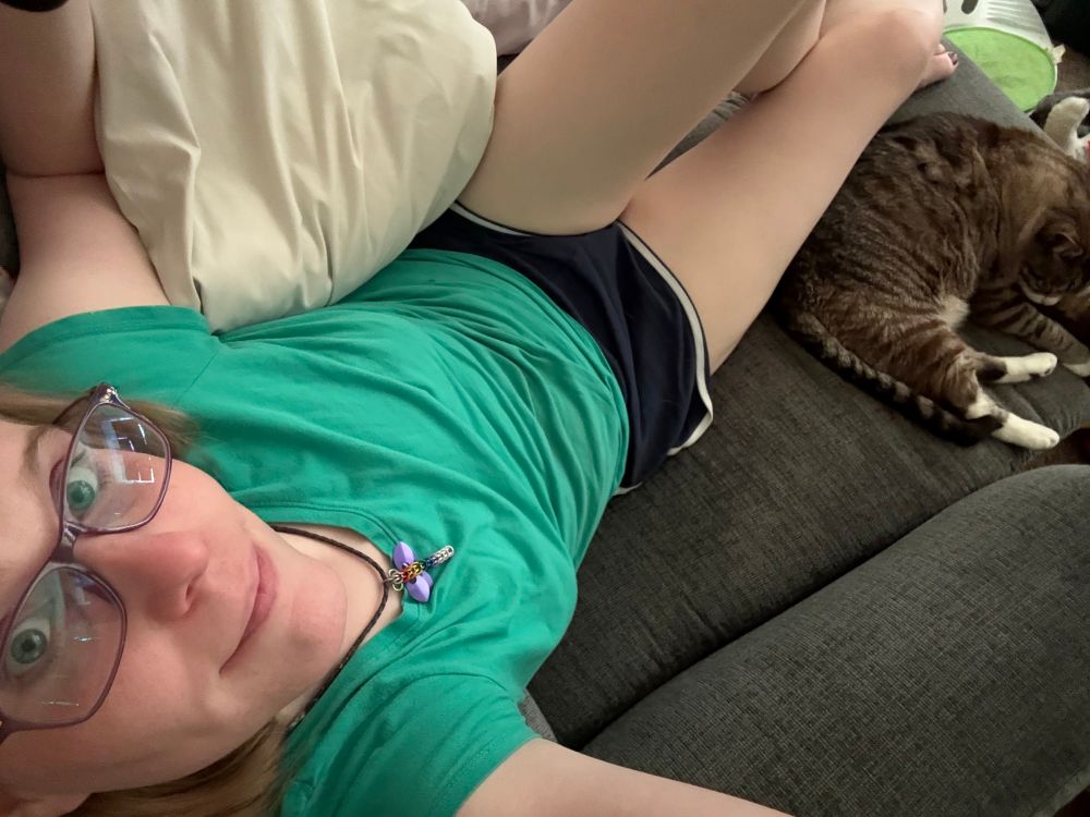 A high angle selfie of a woman wearing a green t shirt and black shorts smiling at the camera with a sleeping cat beside her on the extended footrest of the recliner they are both on