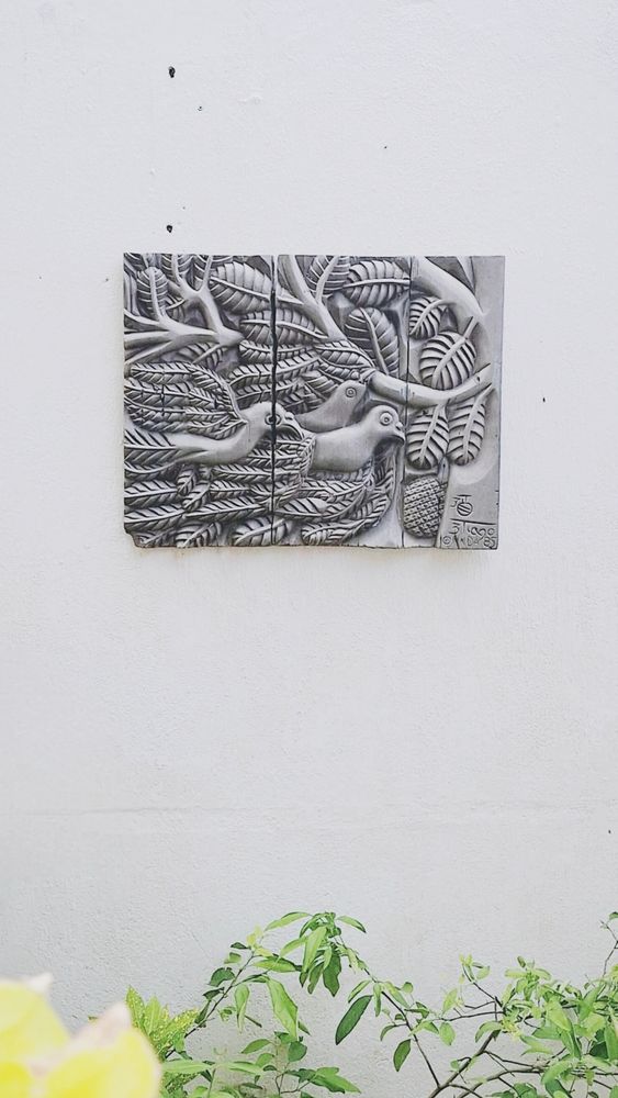 Uma placa decorativa em relevo pendurada em uma parede branca. A placa é composta por três painéis verticais e apresenta uma cena naturalista esculpida, com três pássaros aninhados em meio a folhagens e galhos. A escultura é detalhada e parece ser feita de madeira ou pedra. A parte inferior da imagem mostra a vegetação, com folhas verdes.