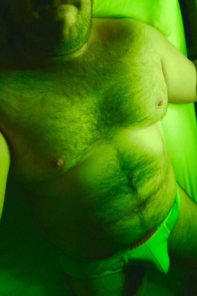 A chubby, hairy man laying in bed showing off his body and bulge in his Aussiebum briefs. 
He is bathed in lime green light