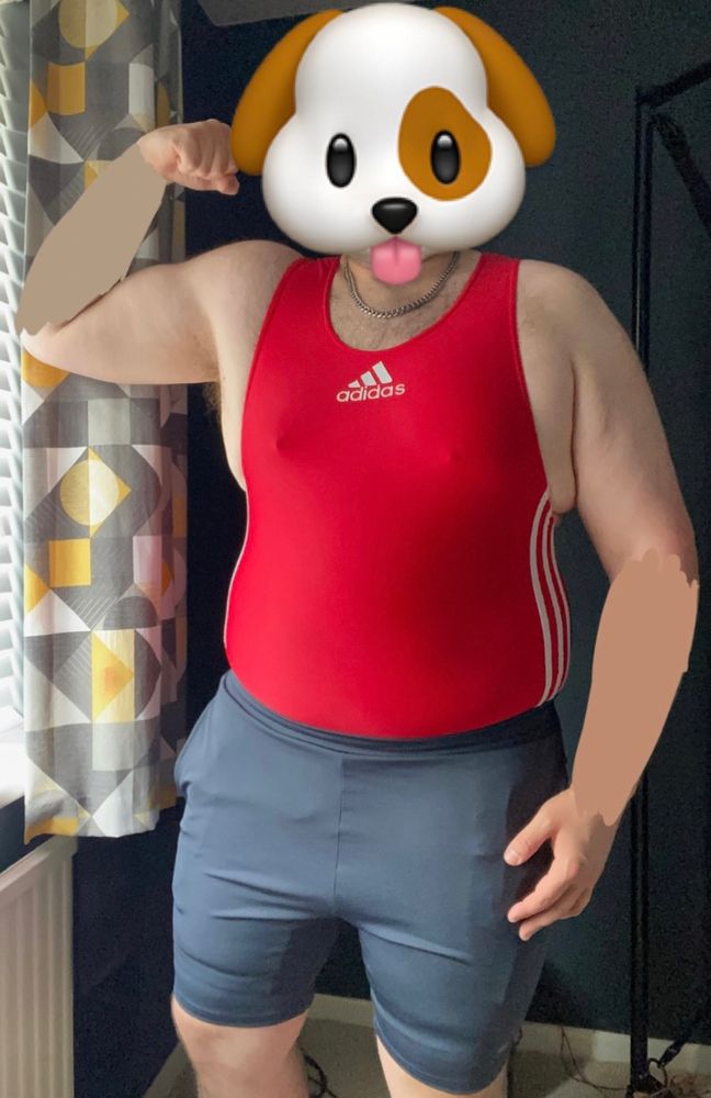 A picture of me in a dark grey room. I'm in grey sorts shorts with a red Adidas singlet underneath as I flex my right bicep