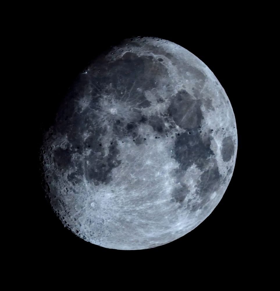 Small-telescope view of nearly full Moon with space station barely resolved as a line of about 18 small black dots.