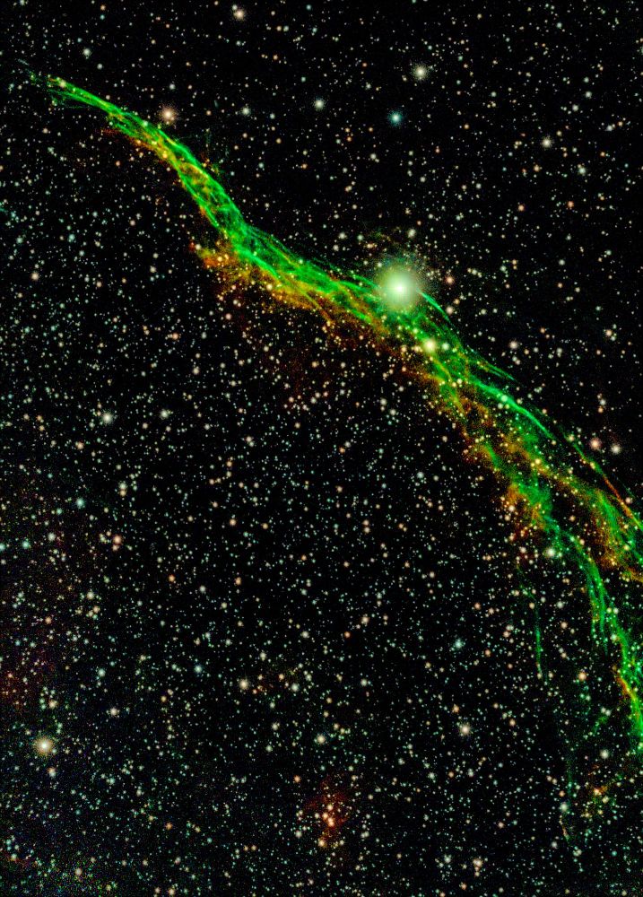 Amateur photo of green and orange filaments of nebula against a background crowded with stars.