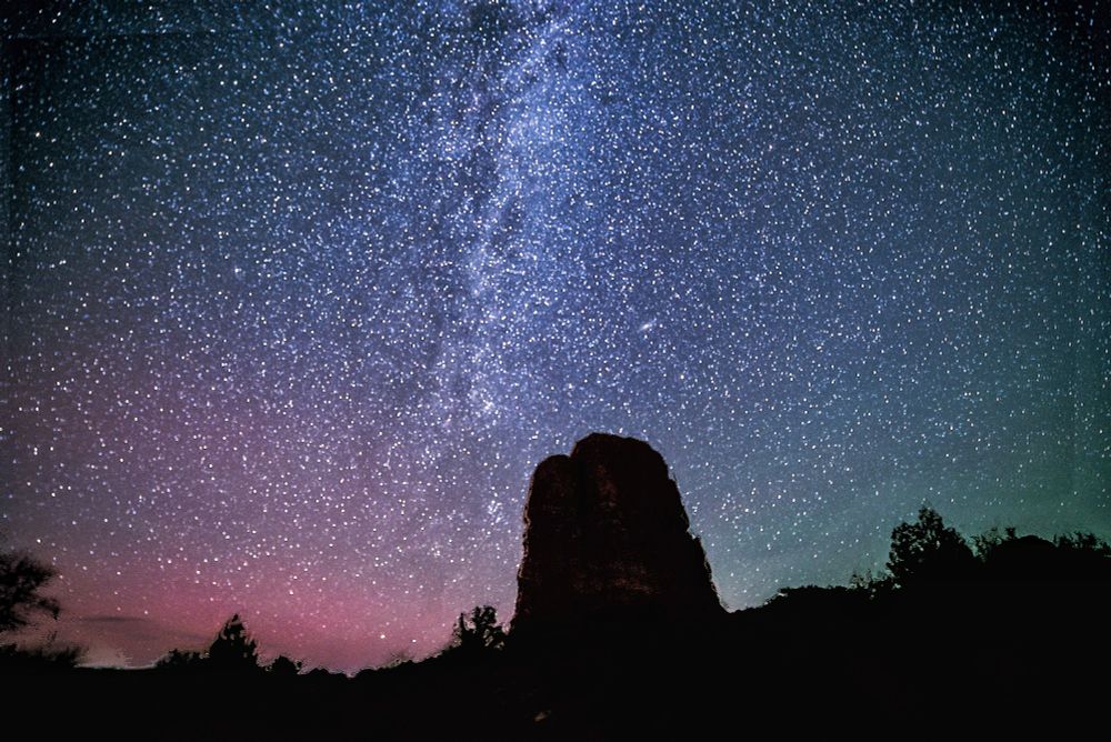 Nighttime photo with the black silhouette of a desert rock formation against the Milky Way and a starry sky tinted red toward the horizon.