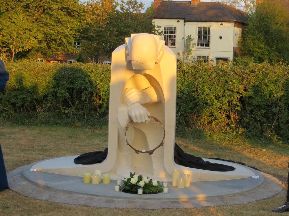 A view from the front of the kneeling figure in the setting sun. From this angle, it is possible to see that he is on one knee, holding a simple crown in his hand as he prays. Black cloth in a heap behind indicates that the statue has just been revealed.
