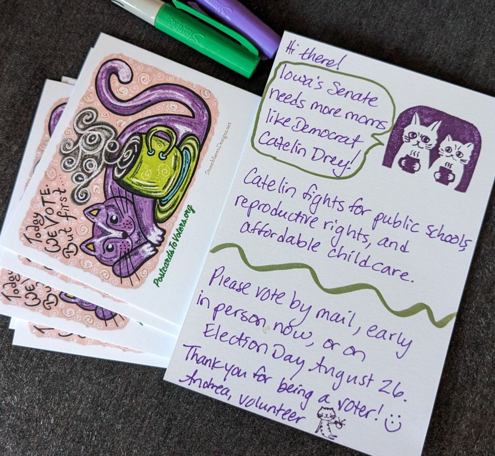 Two postcards laying on a dark surface with a purple and a green pen. One card shows the stamped side with a cartoon drawing of a happy cat curled around a steaming mug of coffee or tea (probably). Text above the cat says "Today we vote. But first...." The second card shows the handwritten side in purple ink saying "Hi there! Iowa's Senate needs more moms like Democrat Catelin Drey. Catelin fights for public schools, reproductive rights, and affordable childcare. Please vote by mail, early in person now or on Election Day, Aug. 26. Thank you for being a voter! Andrea, volunteer" There is a cartoon on the right side of two cats sitting next to each other holding mugs with steaming liquid inside.