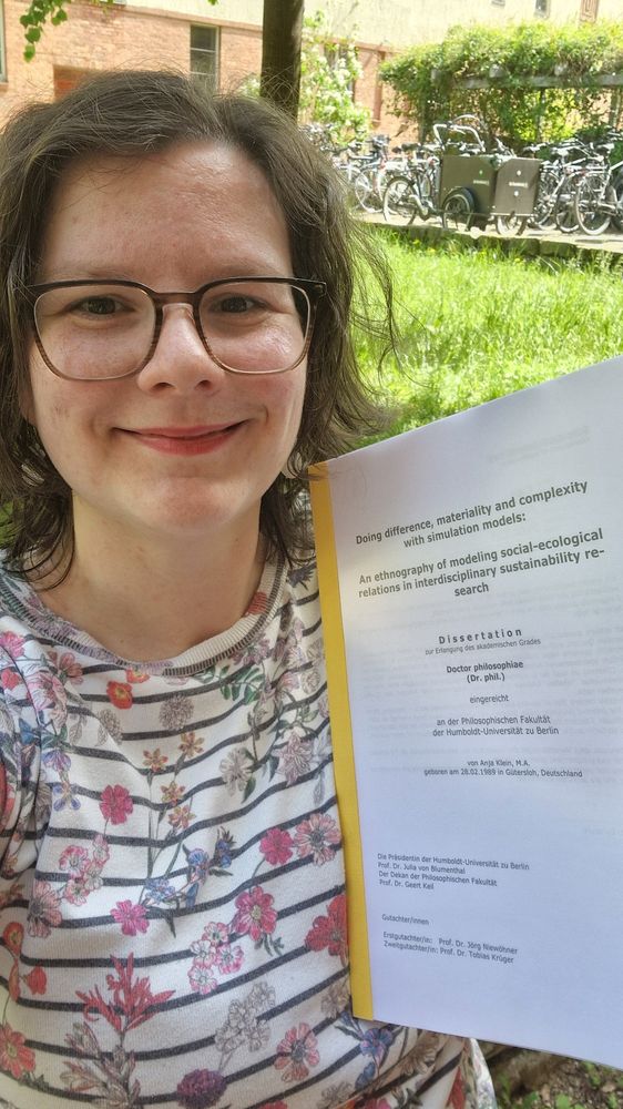 Selfie lf myself, smiling, holding my printed dissertation on one arm, the title reads "Doing difference, materiality and complexity with simulation models. An ethnography pf modeling social ecological relations in interdisciplinary sustainability research "