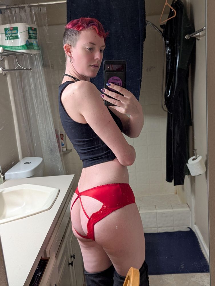 Delia takes a mirror selfie over their shoulder wearing a black crop top and bright red open back panties that frame their ass. Their jeans are pulled down for the reveal