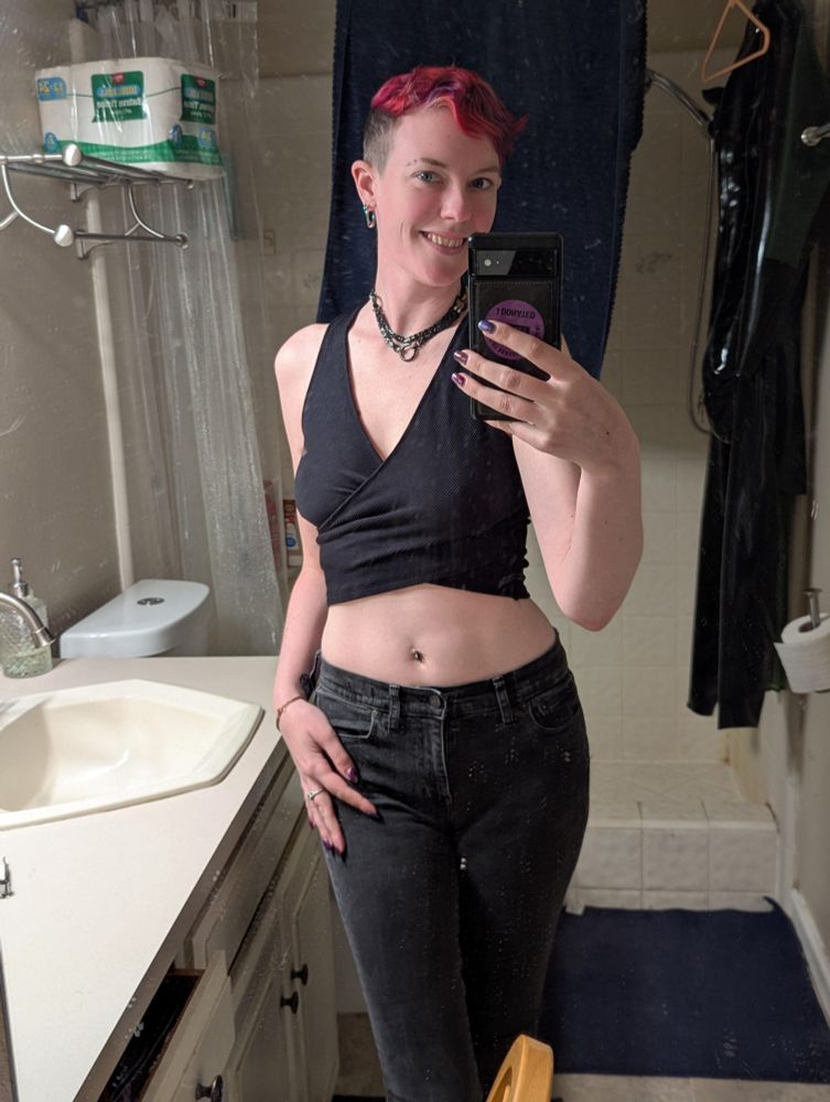 Delia stands smiling in a mirror selfie wearing a black crop top and dark gray skinny jeans 
