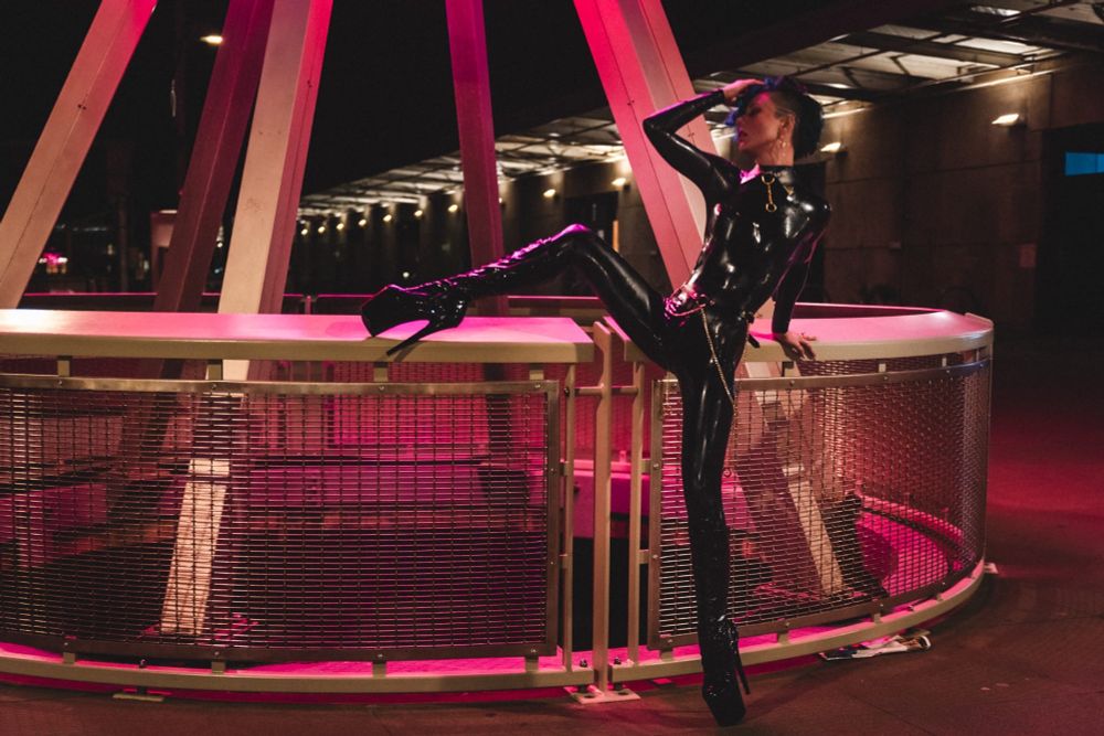 Mixtress Ryder puts one leg up on a railing in red light, wearing a full latex catsuit with bondage details and tall, patent platform heels