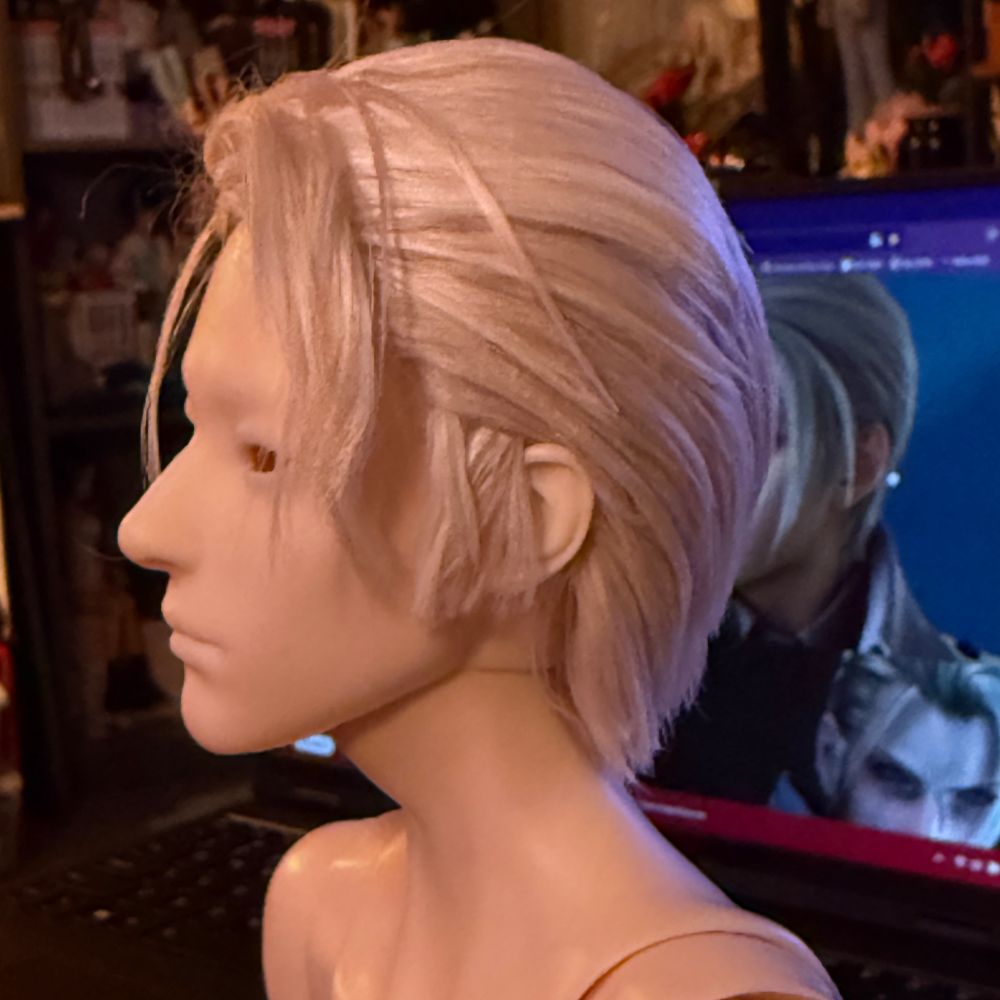 Unpainted and eyeless doll with blond wig styled in swept back style to look like Rufus Shinra from Final Fantasy 7. Profile view.