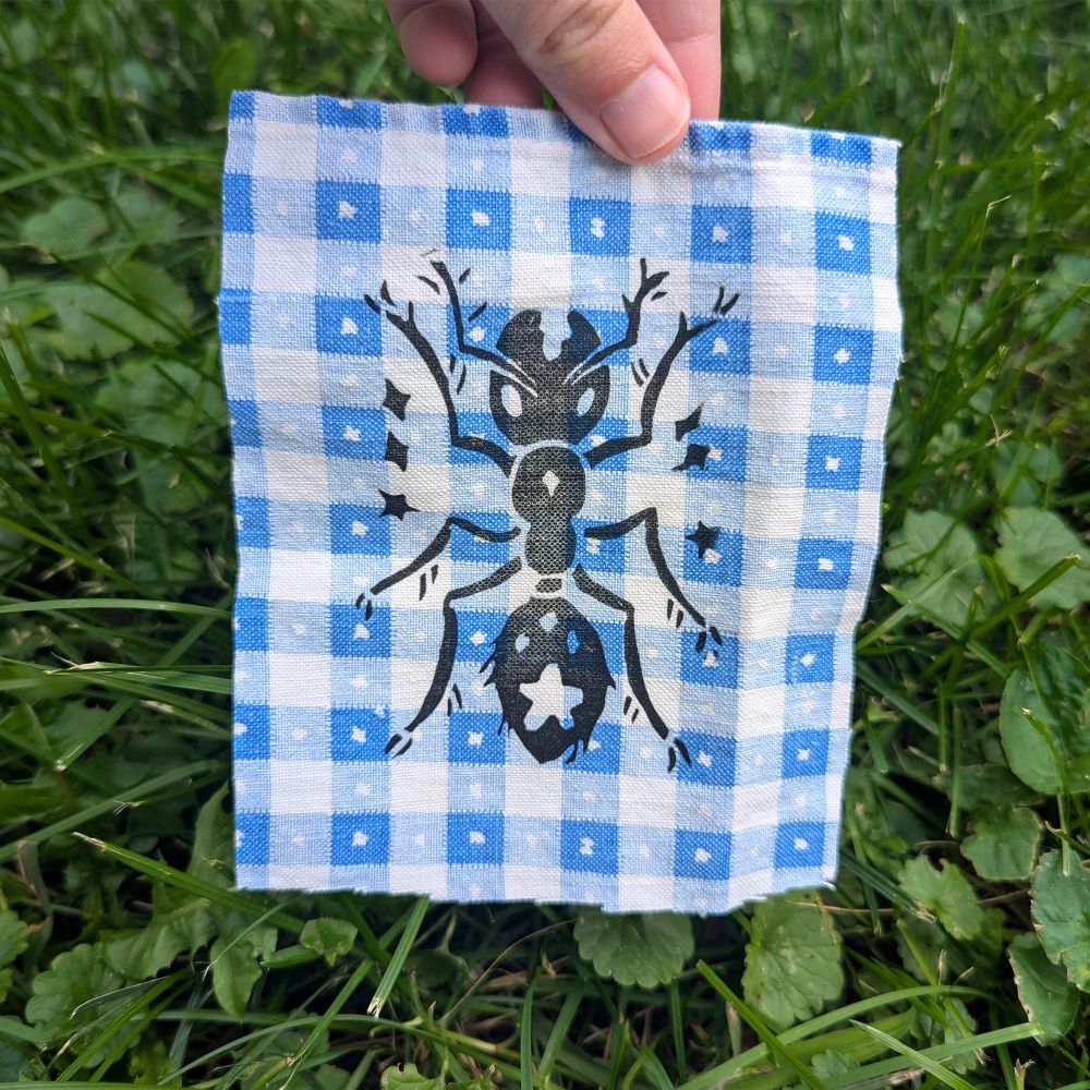A linocut print of an ant on a checkered fabric, reminiscent of a picnic blanket.