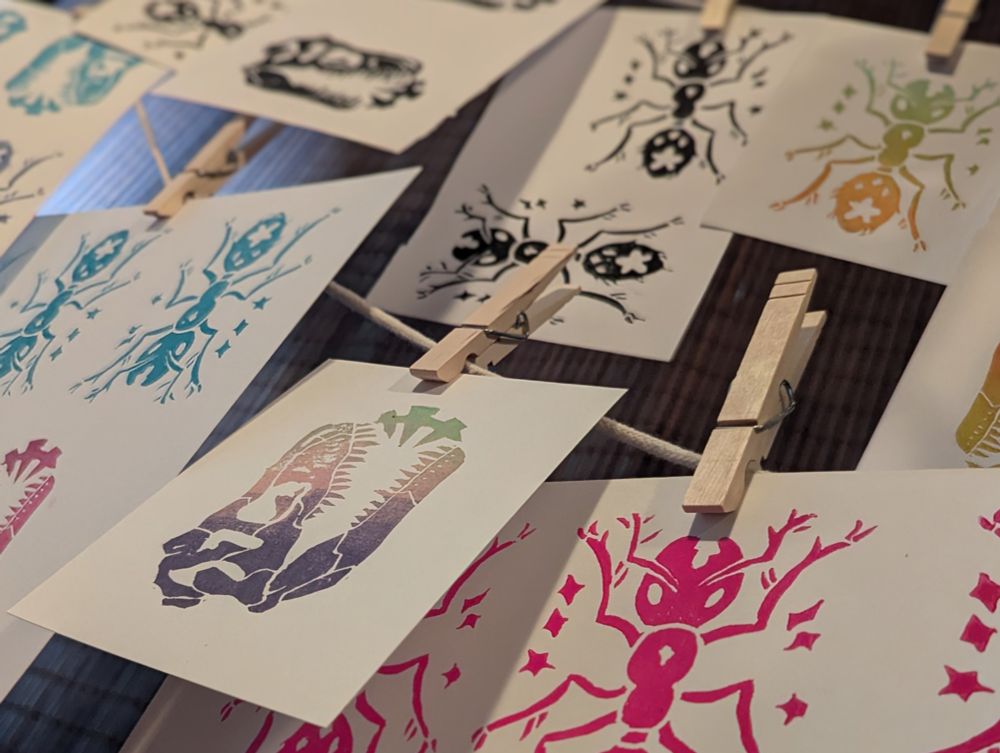 Photo of multiple multi-color lino-prints of a T-Rex skull and an ant hanging on a line drying.