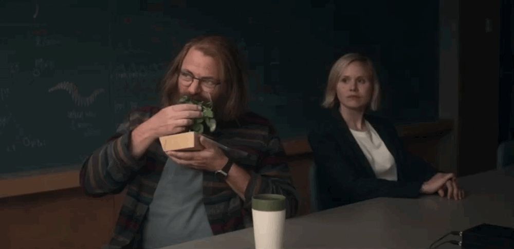 dois personagens da série DEVS. um homem e uma mulher sentados à uma mesa. o homem comendo uma salada de folhas de forma voraz e a mulher apenas observando.