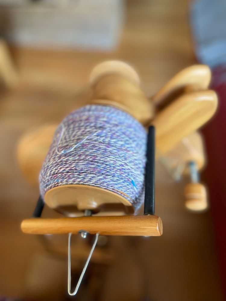Closeup of a spinning wheel flyer with a bobbin full of two-ply woolen yarn in purple and natural white. 
