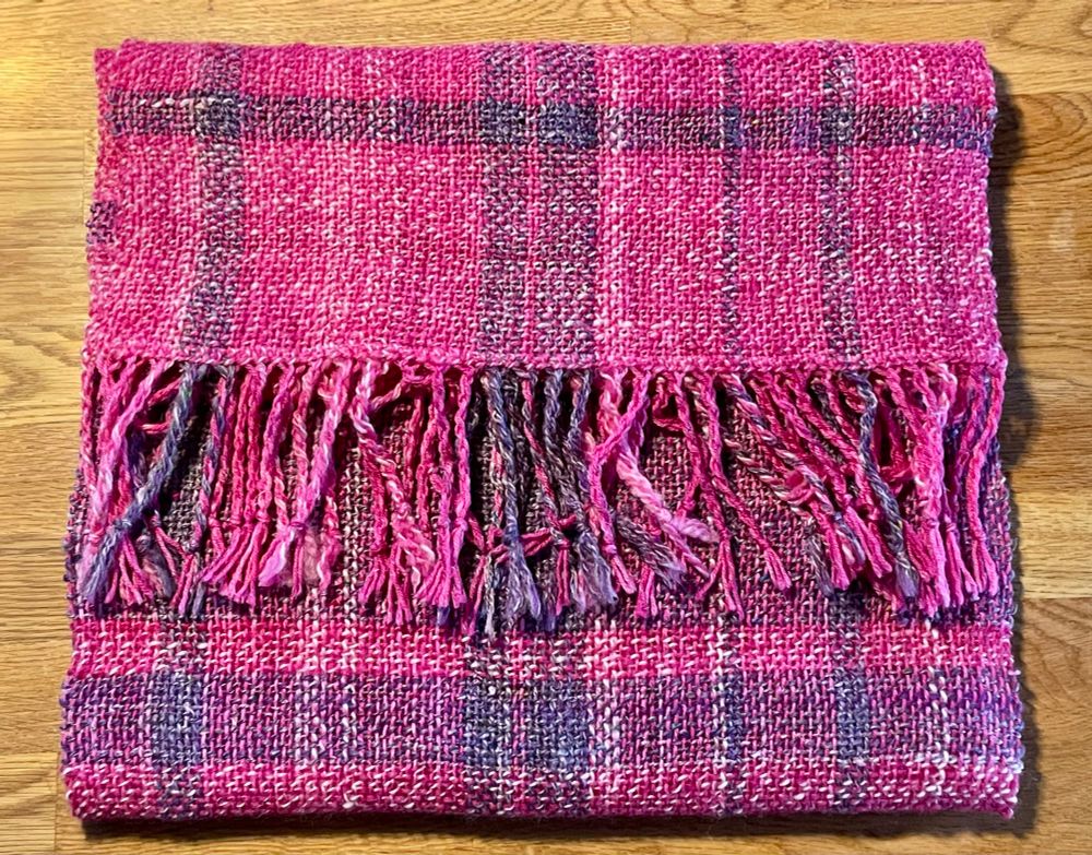 Scarf with a fringe, handwoven, pinks and purples. Folded on a table. 