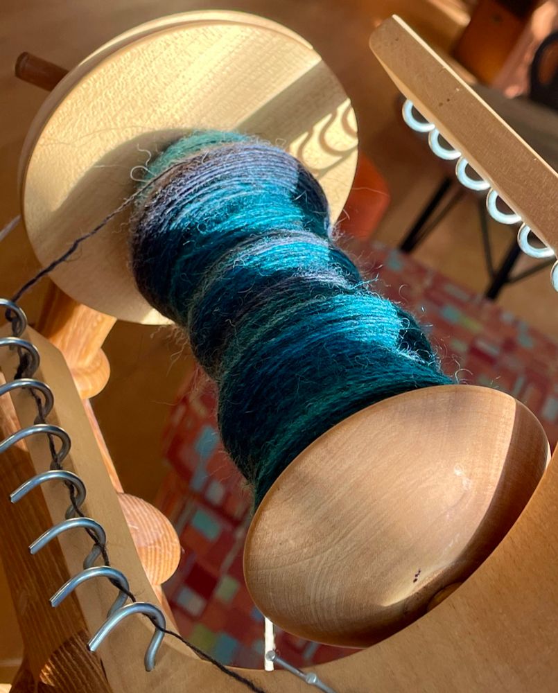 Closeup of the bobbin of a spinning wheel with yarn on it in blue/black/teal colours. 