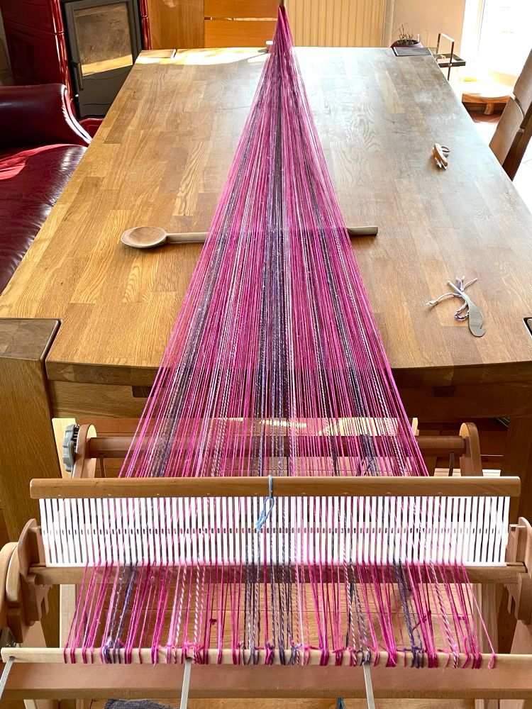 A colourful warp stretched out from the loom across the table to the peg at the end for direct warping. Mostly shades of pink with some purple.