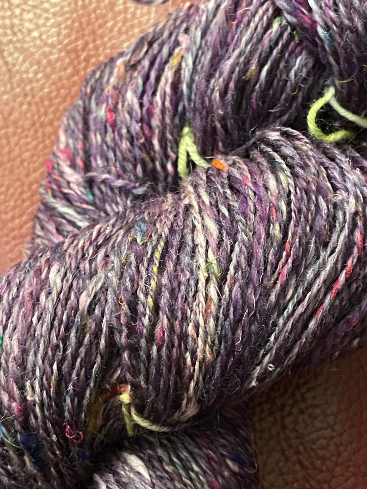 Closeup of a skein of yarn. Two-ply, mainly purple with flecks of colour and little bumps from the silk. 